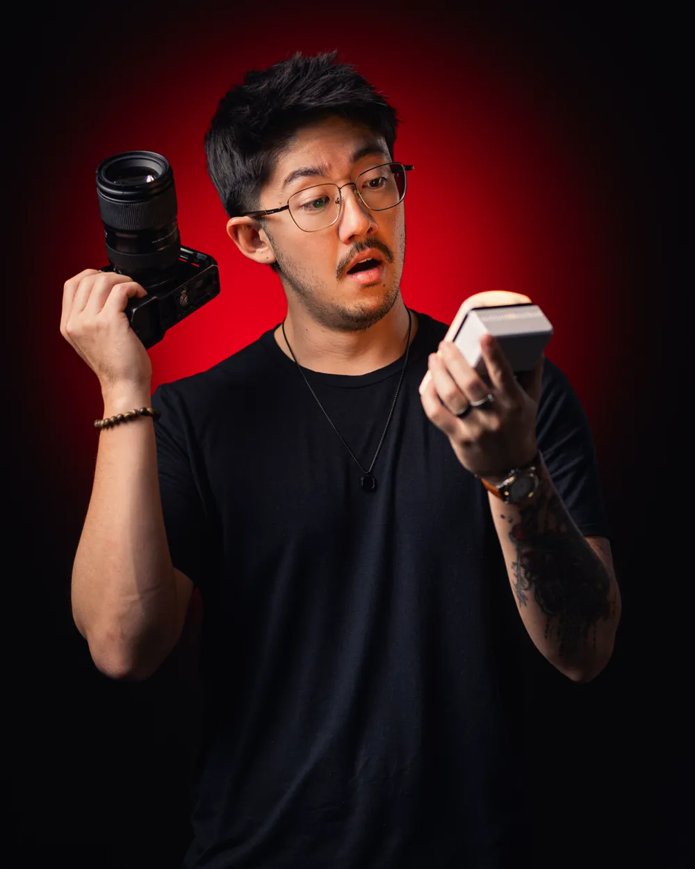 Thiago Kai em fundo vermelho, com uma câmera numa mão e uma luz de LED na outra, olhando a luz surpreso.